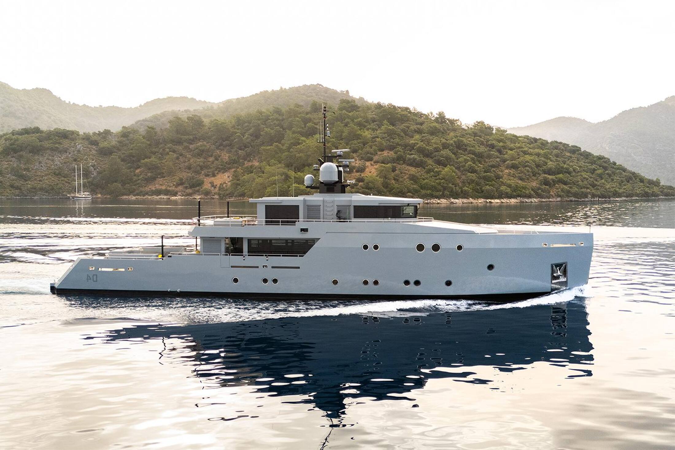 A grey-hulled explorer yacht under way in a calm bay lined with pine-covered hills