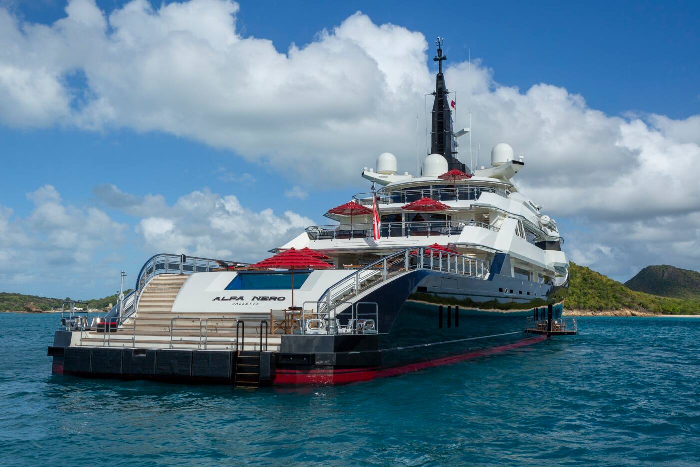 ALFA NERO at anchor off a green island, her cascading aft steps and beach club open to the water