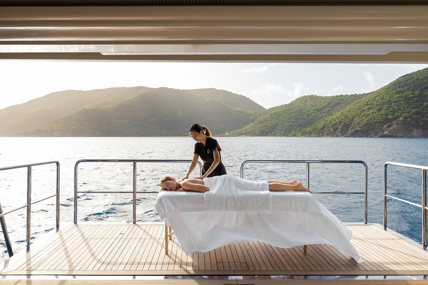A guest receiving a massage on the shaded aft deck of CARINTHIA VII, overlooking a green headland