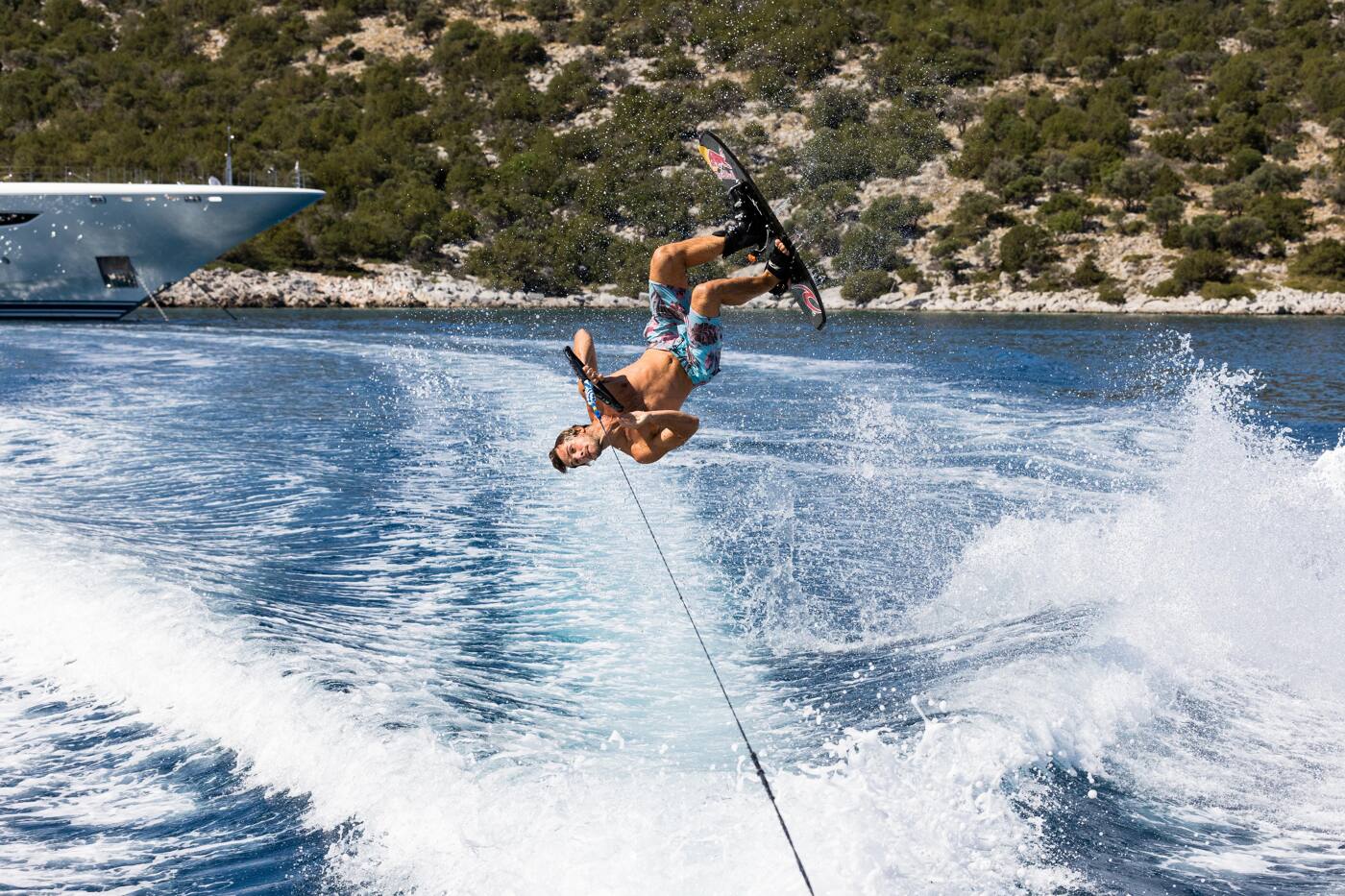 A wakeboarder mid-flip in the wake, with a superyacht anchored against a pine-covered shore